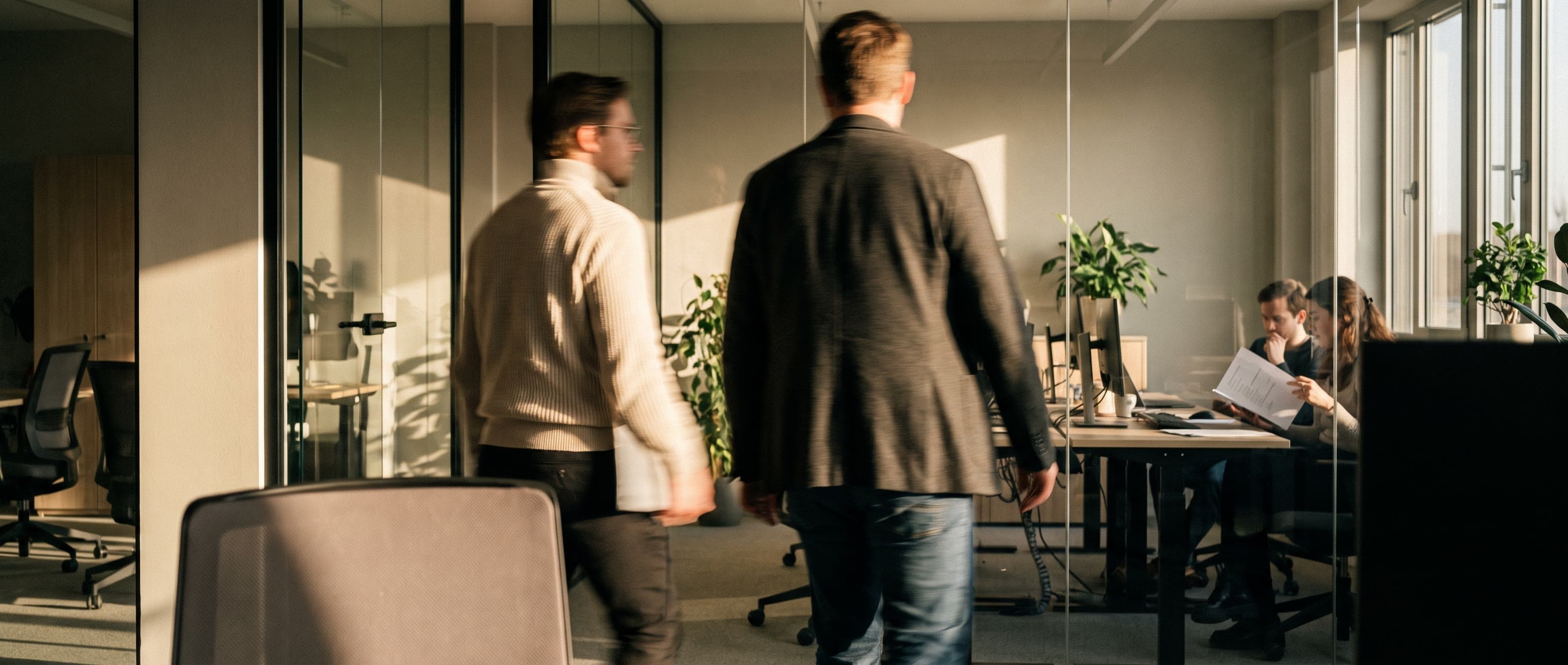 Two colleagues crossing a working office floor at speed, motion-blurred against the sharp room behind them, late window light raking in low from one side.